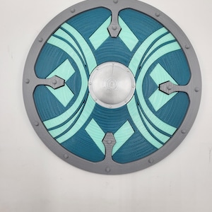 May include: A circular shield featuring a central silver-colored boss, surrounded by a dark teal and light mint green geometric pattern. The outer rim is grey with evenly spaced circular studs, creating a structured, layered appearance.