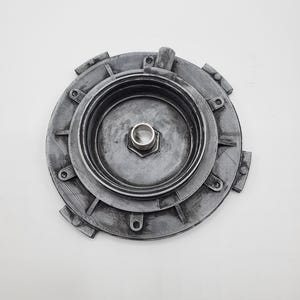 Puede incluir: Un componente mecánico circular gris con una tuerca hexagonal central y múltiples capas circundantes. El componente tiene una superficie texturizada y varias pestañas que sobresalen alrededor del perímetro. El objeto está sobre un fondo blanco liso.