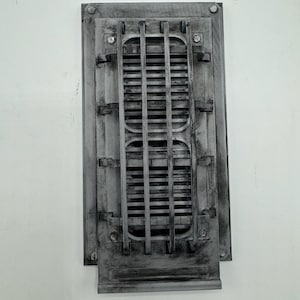 Op de afbeelding: Een rechthoekige, donkergrijze wandventilatiekap met een verweerde uitstraling, voorzien van een centraal roosterpatroon met verticale en horizontale staven. Het frame heeft een versleten afwerking met zichtbare schroefachtige details.