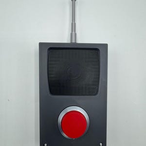 May include: A rectangular dark gray controller featuring a large, circular red button centered in a silver metallic ring. Above the button is a recessed black square panel with a textured surface. A thin, silver, cylindrical antenna extends vertically from the top edge of the device.