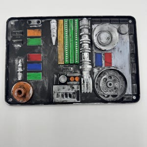 Könnte beinhalten: Eine rechteckige schwarze Platte mit einer Vielzahl erhabener mechanischer Komponenten, darunter silberne Metallzylinder, grüne und goldene Stäbe sowie kleine rote, blaue und orangefarbene rechteckige Akzente mit einem verwitterten, industriellen Finish.