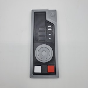 Peut inclure: Un panneau de contrôle rectangulaire avec un cadre gris et une section centrale noire. Il comprend un cadran circulaire gris, deux petits boutons rectangulaires blancs, deux petits boutons circulaires blancs, un bouton carré blanc et un bouton carré rouge.