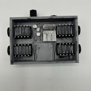 May include: A rectangular grey plastic enclosure featuring four black ribbed components, three silver cylindrical elements, and three silver rectangular blocks. The device includes black knobs on the sides and top, with a weathered, distressed finish.