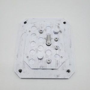 May include: A white, rectangular mechanical component with circular and hexagonal openings. It features a central metal rod and is secured with bolts. The surface has black smudges for texture.