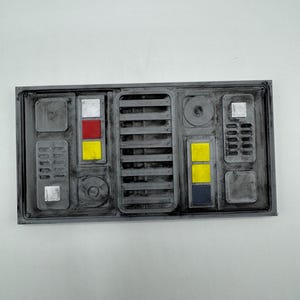May include: A rectangular grey 3D-printed panel featuring a central vertical vent, circular dials, and recessed square sections. The panel includes small, flat, coloured squares in red, yellow, and dark blue, along with silver-toned accents.