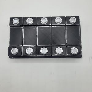 Puede incluir: Un componente rectangular negro con diez puertos hexagonales de borde plateado dispuestos en dos filas de cinco. La sección central consta de cinco paneles rectangulares negros verticales, con los puertos situados en los bordes superior e inferior.