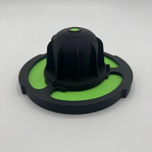 Puede incluir: Un componente negro impreso en 3D con forma de cúpula y nervaduras verticales, centrado sobre una base circular plana. La base presenta un inserto circular verde brillante con dos recortes simétricos, enmarcado por un anillo exterior negro.