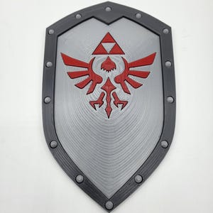 Puede incluir: Un escudo con base puntiaguda y borde gris oscuro con remaches circulares. El centro es gris claro con un patrón texturizado de círculos concéntricos y un emblema rojo que representa un ave estilizada y un símbolo triangular.