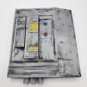 May include: A rectangular grey panel with a weathered, metallic finish featuring raised sections, two yellow vents, a blue circular button, and a red circular button. The surface has visible 3D printed texture lines and three cylindrical protrusions at the base.