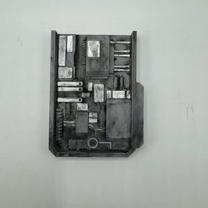 Puede incluir: Una placa de pared rectangular de color gris oscuro con un complejo patrón de relieve mecánico. La superficie muestra varias formas geométricas, incluyendo rectángulos, cuadrados y un anillo circular, con acabados en plata metálica.