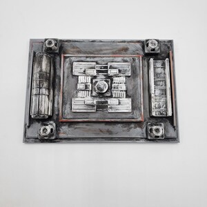 May include: A rectangular, gray, metallic-looking wall art piece with a complex, raised design. The central area features a circular element surrounded by geometric shapes. The corners have cylindrical and square elements. The piece has a textured appearance.