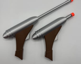 May include: Two silver and brown toy blasters with long barrels and orange tips. Each blaster features a metallic silver main body, a brown handle, and a silver trigger. The blasters are positioned side by side, with one having a longer barrel.