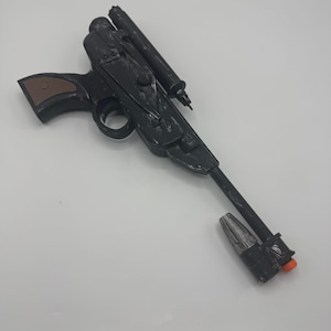 以下が含まれることがあります： 長い銃身、上部に取り付けられたスコープ、茶色のグリップを備えた黒いおもちゃのブラスター。銃身には銀色のマズルアタッチメントと小さなオレンジ色の先端が付いています。全体的なデザインには、成形された機械的なディテールとトリガーガードが含まれています。