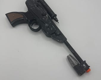 May include: A black toy blaster with a long barrel, a top-mounted scope, and a brown grip. The barrel features a silver-colored muzzle attachment and a small orange tip. The overall design includes molded mechanical details and a trigger guard.
