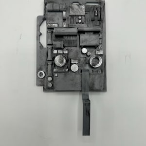 Peut inclure: Un panneau mécanique gris rectangulaire présentant un agencement complexe de formes géométriques en relief, de composants circulaires et de piliers verticaux. La surface a une finition métallique patinée avec un bras saillant vers le bas.