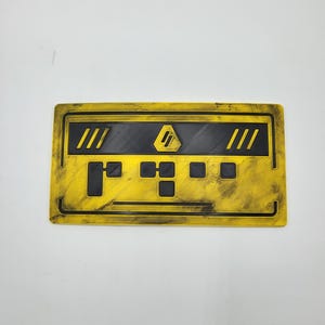 Könnte beinhalten: Eine rechteckige gelb-schwarze Plakette mit verwitterter, abgenutzter Oberfläche. Das Design umfasst ein zentrales schwarzes horizontales Band mit einem gelben sechseckigen Symbol, flankiert von drei diagonalen Linien auf jeder Seite und schwarzen quadratischen Ausschnitten.