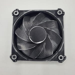 May include: A square black cooling fan with a circular center and curved blades. The frame features four mounting holes at the corners, each reinforced with a metallic ring. The surface of the fan shows a textured, layered finish consistent with 3D printing, with visible lines across the blades and the outer housing.