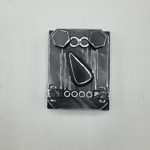 May include: A rectangular black object with a textured surface featuring two hexagonal shapes at the top, a central teardrop-shaped protrusion, and a row of four circular rings positioned above two small rectangular latches at the bottom edge.