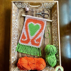 May include: A crocheted wall hanging with a heart design in shades of green, orange, and pink. The hanging has green fringe and is displayed in a wooden tray with balls of orange and green yarn.