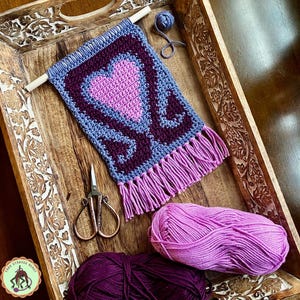 May include: A crocheted wall hanging with a heart design in shades of purple, pink, and blue. The hanging is displayed in a wooden tray with yarn balls and scissors.