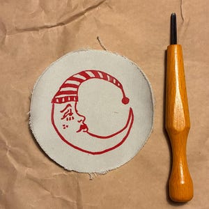 May include: A circular patch with a red smiling moon design wearing a striped cap. The patch is on a light beige fabric. A wooden-handled tool with a black tip is next to the patch.