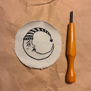 May include: A circular patch with a black and white crescent moon design, next to a wooden-handled tool. The moon has a face and a striped hat. The tool has a metal tip and a varnished wooden handle.