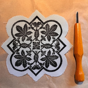 May include: A white fabric square with a black floral design and a wooden carving tool. The fabric has a scalloped edge and features a symmetrical pattern of leaves and flowers. The carving tool has a wooden handle and a metal blade.