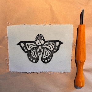 May include: A black butterfly design printed on a light gray fabric square with frayed edges. A wooden carving tool with a metal blade is positioned to the right of the fabric. The background is a light brown surface.
