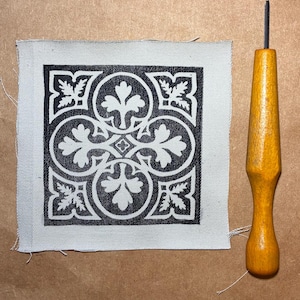 May include: A square piece of fabric with a black and white floral design, next to a wooden tool with a metal tip. The fabric has a repeating pattern of circles and leaves. The tool has a yellow wooden handle.