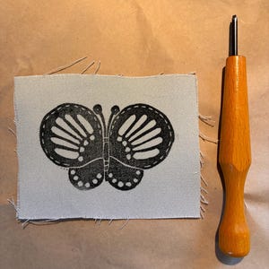 May include: A black butterfly design printed on a square of light gray fabric. The butterfly has detailed wing patterns. A wooden-handled punch needle tool is positioned next to the fabric, suggesting a craft project.