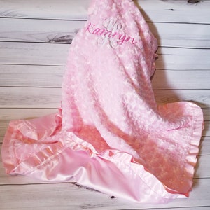 May include: A pink baby blanket with a satin border and the name "Kamryn" embroidered on it.