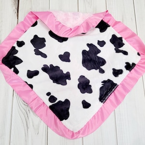 May include: A soft, white and black cow print baby blanket with a pink satin trim. The blanket is made of plush fabric and is perfect for cuddling.