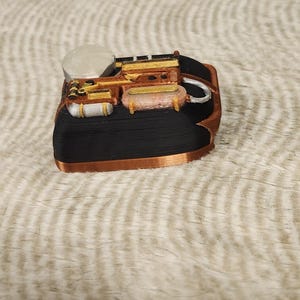 May include: A 3D-printed miniature steampunk-style train engine. The model features a black base with copper-colored trim and detailed components in gold, brown, and white. The design includes a smokestack, boiler, and various mechanical elements.