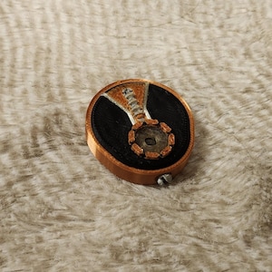 Peut inclure: Un objet rond de couleur cuivrée avec un centre noir et un motif détaillé en forme de roue dentée. L'objet a une vis sur le côté et est posé sur un fond doux et texturé de couleur claire.