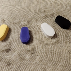 May include: Four colorful, oval-shaped objects in yellow, blue, white, and black are arranged on a textured, tan surface. The objects have a rounded top and a flat bottom, suggesting a potential functional design.