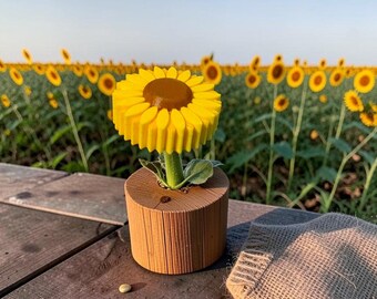 Può includere: Un girasole stampato in 3D con petali giallo brillante e un centro marrone, inserito in una base cilindrica marrone con scanalature verticali. Piccoli semi verde chiaro sono sparsi sulla superficie in legno accanto a un pezzo di iuta.