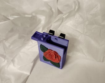 Può includere: Una piccola scatola viola con un coperchio incernierato e un design a rosa rossa. La scatola è fatta di plastica e ha una cerniera nera.