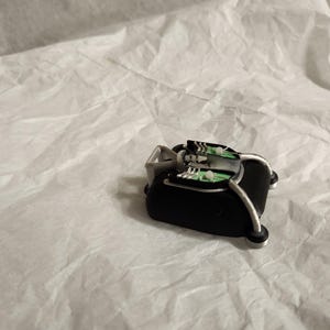 May include: A small, black, stylized object with silver accents and a green circuit board design. The object is resting on a crumpled white surface, creating a contrast in textures and colors. The design suggests a technological or miniature device.