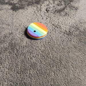 May include: A small, round, rainbow-colored wooden button with a hole in the center. The button is made of several layers of wood, each painted a different color of the rainbow. The button is lying on a gray, textured surface.