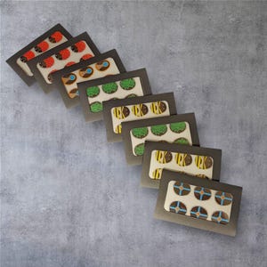 May include: A collection of seven small, dark gray boxes, each with a clear window revealing a different design of decorated cookies. The cookies feature various designs, including ladybugs, shields, and bees, with a neutral background.