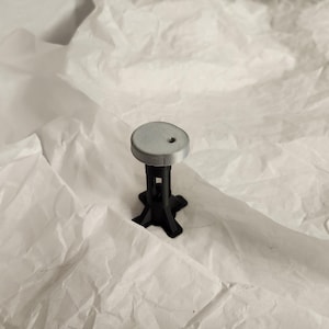 Op de afbeelding: Een kleine, ronde grijze kruk met een zwarte basis. De kruk is gemaakt van plastic en heeft een eenvoudig ontwerp.