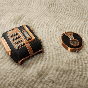 Puede incluir: Dos accesorios de estilo steampunk. Uno es una caja cuadrada negra y cobriza con detalles intrincados. El otro es un medallón ovalado cobrizo con un centro negro y detalles. Ambos artículos parecen estar impresos en 3D.