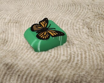 Può includere: Un oggetto verde di forma quadrata con un disegno a farfalla. La farfalla ha ali nere e gialle. L'oggetto è appoggiato su una superficie bianca e strutturata.