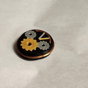 May include: A small, round, black and copper-colored object with three gears on top. The gears are silver, gold, and copper.