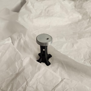 Op de afbeelding: Een klein, grijs en zwart 3D-geprint object dat lijkt op een miniatuurkruk. De kruk heeft een cirkelvormige grijze bovenkant met een klein gaatje, ondersteund door een zwarte, geometrische basis. Het object staat tegen een witte, getextureerde achtergrond.