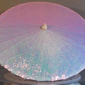 Puede incluir: Un parasol decorativo con un degradado de colores rosa, morado y azul, cubierto de purpurina brillante. Tiene un pequeño mango beige y está sobre una base circular. El diseño es caprichoso y llamativo.