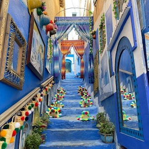 Puede incluir: Un callejón azul vibrante con escaleras, adornado con sombreros y cestas coloridas. Las paredes están pintadas en un llamativo tono azul, con elementos decorativos y plantas en macetas que se suman al encanto de la escena. Las escaleras están bordeadas de sombreros coloridos.