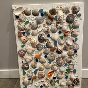 May include: A white canvas with a mosaic of seashells, glass beads, and cork pieces. The seashells are various shapes and sizes, and the glass beads are blue, green, and orange. The cork pieces are brown.