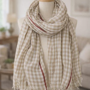 Może przedstawiać: Beżowo-biała szalik w kratę z czerwonym paskiem, prezentowany na manekinie. Szalik ma postrzępioną krawędź i jest udrapowany wokół szyi manekina, prezentując swoją miękką, lekką tkaninę.