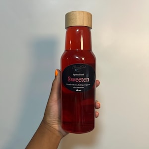 Puede incluir: Un líquido rojo transparente en una botella cilíndrica alta con una tapa de madera marrón claro. La etiqueta ovalada negra presenta el texto Ethereal Soul, Spiritual Bath, Sweeten, Created with love, healing energy and clear intentions, 18 oz.